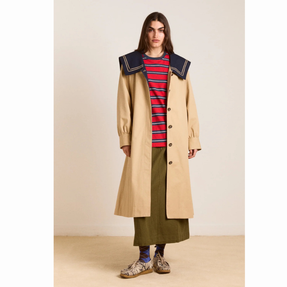Person set forfra iført Damson Madder Ella Trench i beige med stor mørkeblå sailor-krave, brune knapper og lange ærmer. Trenchcoaten bæres åben over en rød- og blåstribet bluse og en lang grøn nederdel.