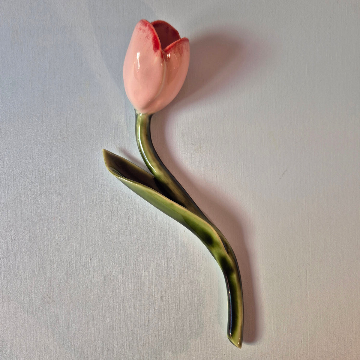 Håndlavet Odd Mud vægkunst formet som en lyserød tulipan med rødlige detaljer, grøn stilk og langt blad på lys baggrund.