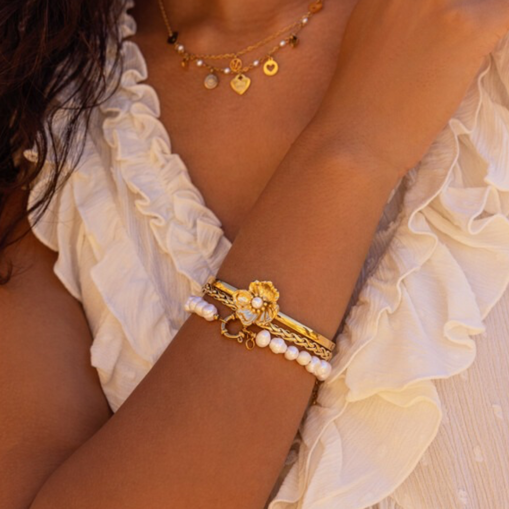 Guld bangle armbånd med en fin dekorativ blomst med perle fra My Jewellery vist på model sammen med et perlearmbånd og guldkæde.