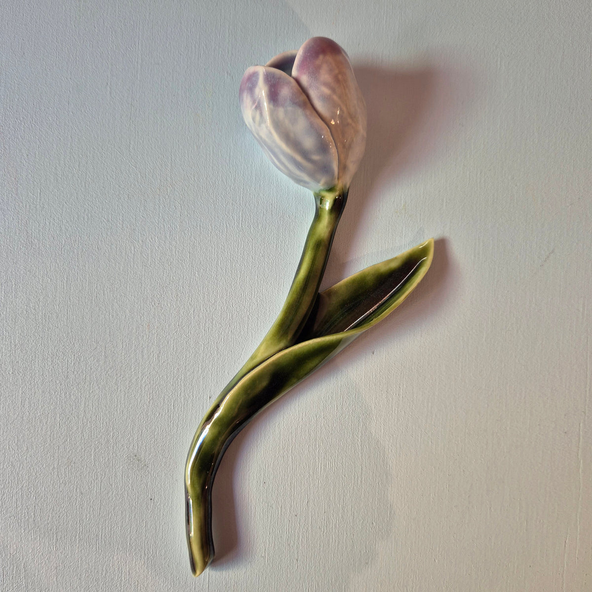 Håndlavet Odd Mud tulipan i keramik med blomst i lilla nuancer, grøn stilk og lange blade på lys baggrund.