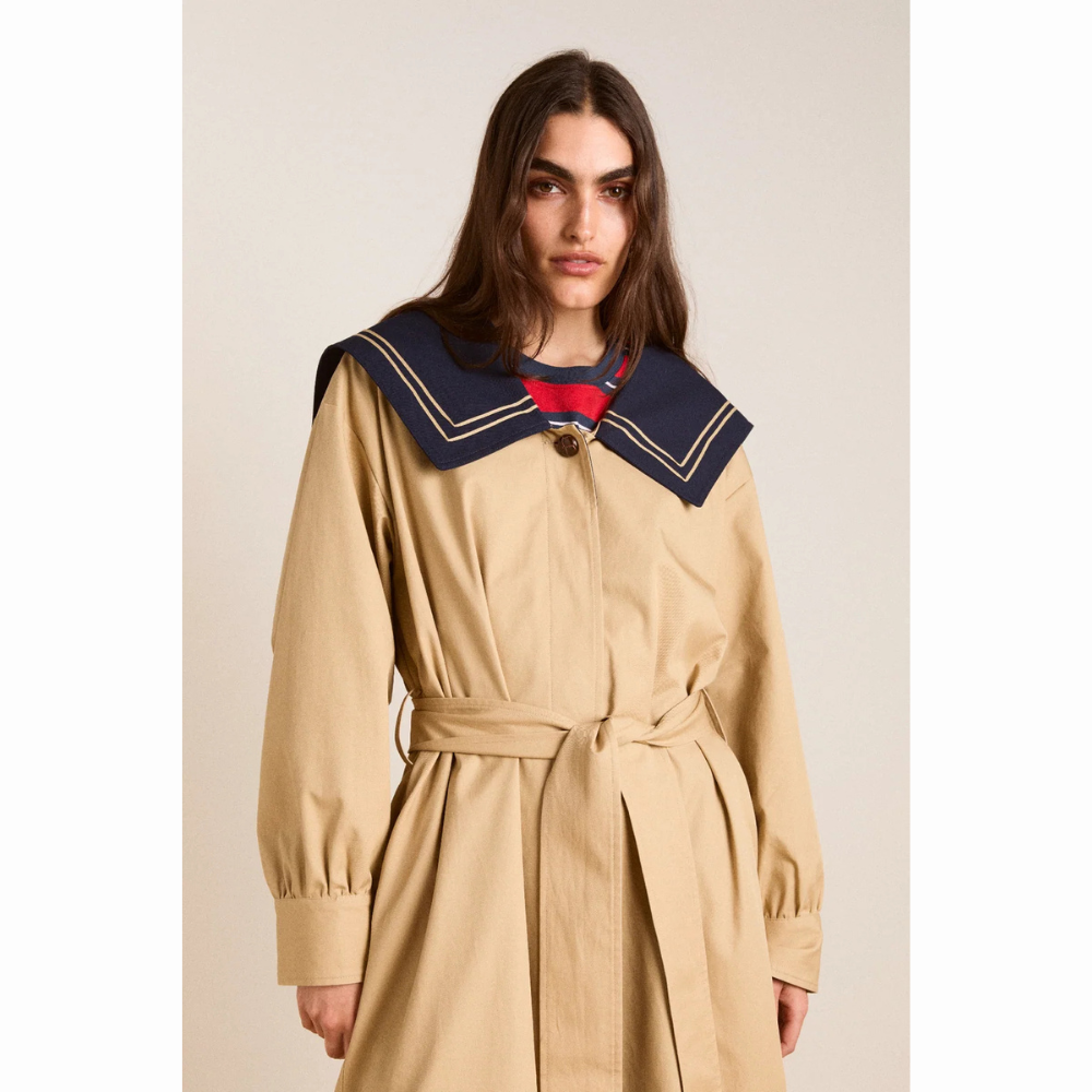 Person set forfra iført Damson Madder Ella Trench i beige med bindebælte i taljen. Den store mørkeblå sailor-krave har beige kantdetaljer og ligger bredt over skuldrene.