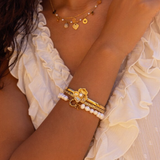 Guld bangle armbånd med en fin dekorativ blomst med perle fra My Jewellery vist på model sammen med et perlearmbånd og guldkæde.