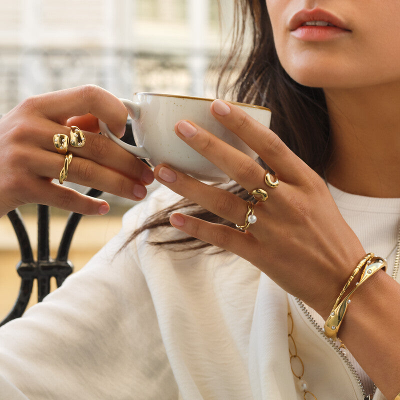 Model bærer guldfarvet ring med perle og bølget design, sammen med flere andre guldsmykker